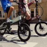 MATE ICON kaufen: Ein mintgrünes und ein rosa Faltrad E-Bike mit Fahrern auf einer Asphaltstraße direkt auf einem aufgemalten Fahrradsymbol.