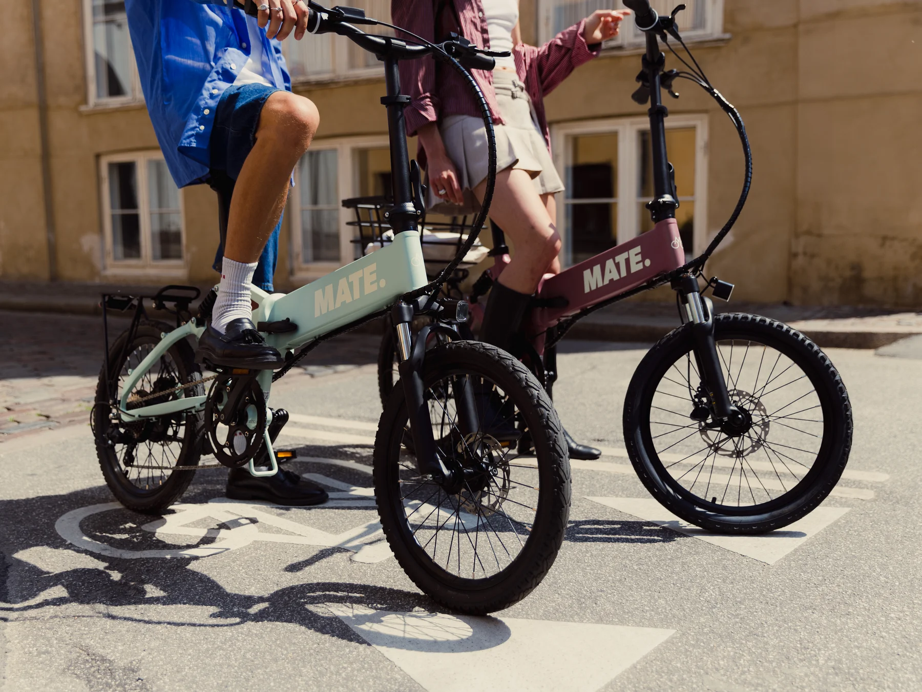 MATE ICON kaufen: Ein mintgrünes und ein rosa Faltrad E-Bike mit Fahrern auf einer Asphaltstraße direkt auf einem aufgemalten Fahrradsymbol.