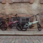 MATE ICON kaufen: Ein rosa Faltrad E-Bike mit schwarzem Fahrradkorb und ein mintgrünes Modell nebeneinander vor einer alten Backsteinwand.