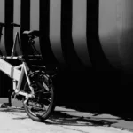 MATE GO kaufen: Helles Faltrad E-Bike auf Betonboden vor einer modernen Wand mit markanten vertikalen Streifen in kontrastreichem Schwarz-Weiß.