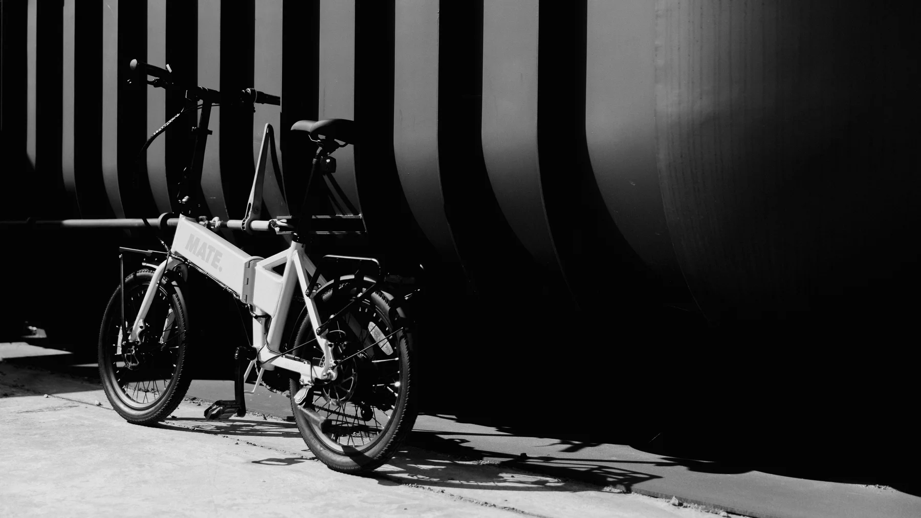 MATE GO kaufen: Helles Faltrad E-Bike auf Betonboden vor einer modernen Wand mit markanten vertikalen Streifen in kontrastreichem Schwarz-Weiß.
