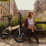 MATE GO kaufen: Beigefarbenes Faltrad E-Bike neben einer jungen Frau auf einer Parkbank inmitten einer blühenden, urbanen Grünanlage.