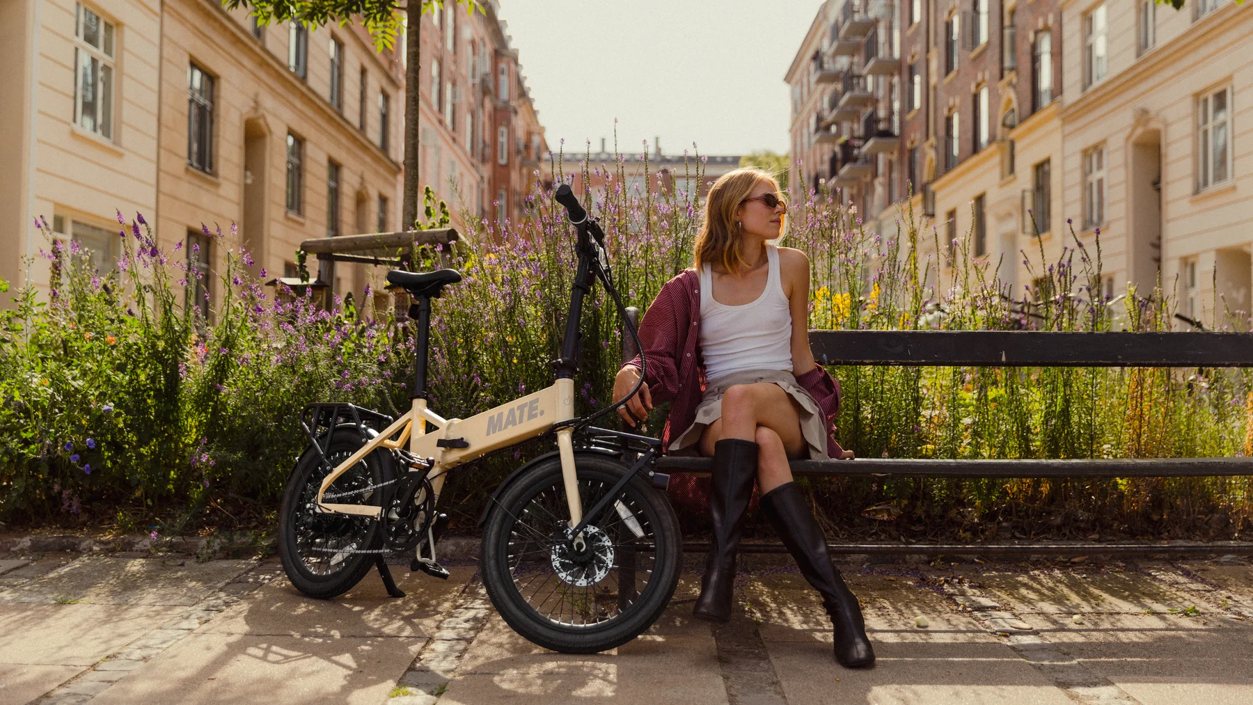 MATE GO kaufen: Beigefarbenes Faltrad E-Bike neben einer jungen Frau auf einer Parkbank inmitten einer blühenden, urbanen Grünanlage.