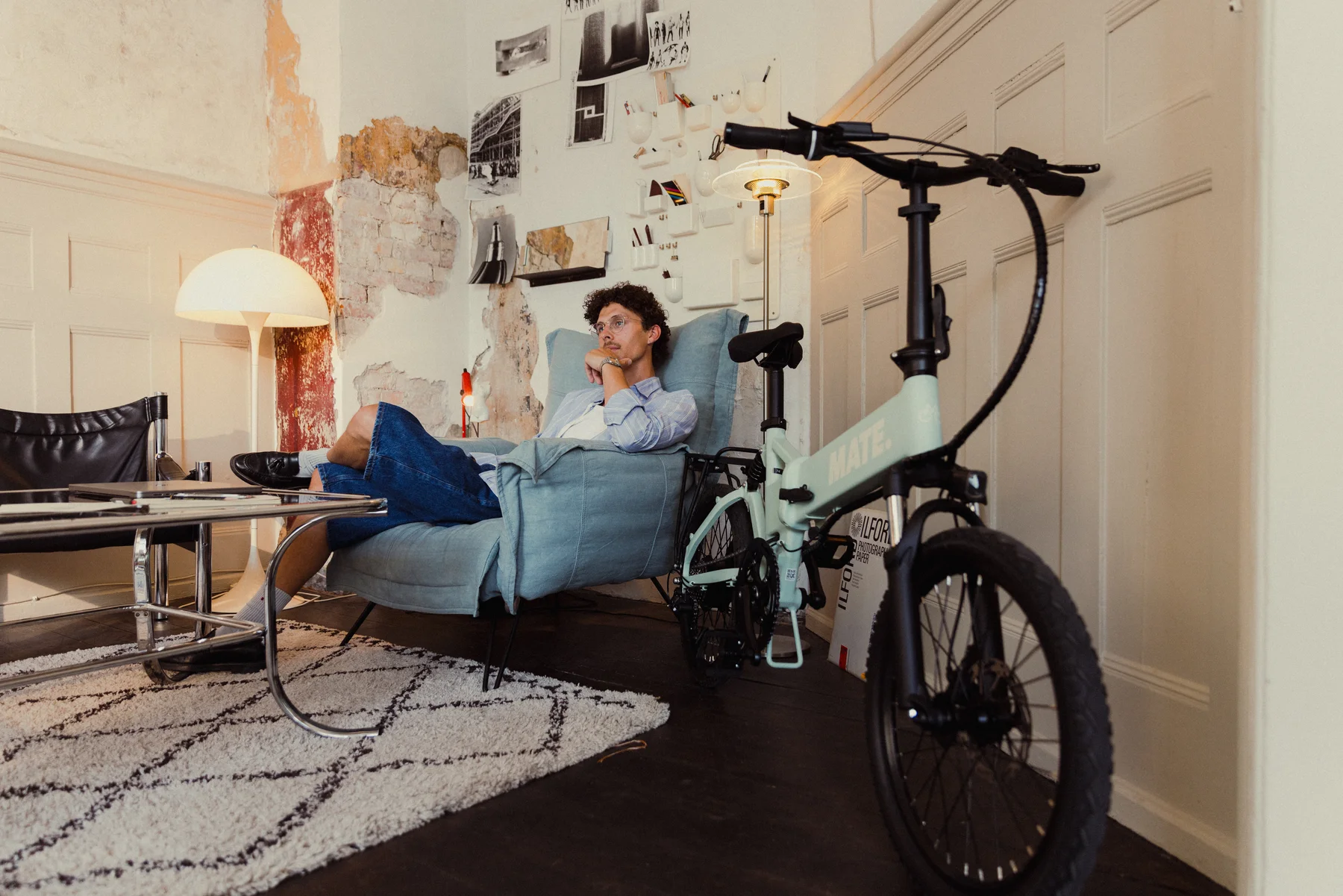 MATE ICON kaufen: Mintgrünes Faltrad E-Bike angelehnt an eine weiße Kassettentür in einer stilvoll eingerichteten Altbau-Wohnung mit einem Mann im Sessel.