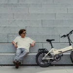 MATE GO kaufen: Beigefarbenes Faltrad E-Bike vor einer großen Steintreppe mit einem entspannt sitzenden jungen Mann mit Sonnenbrille.