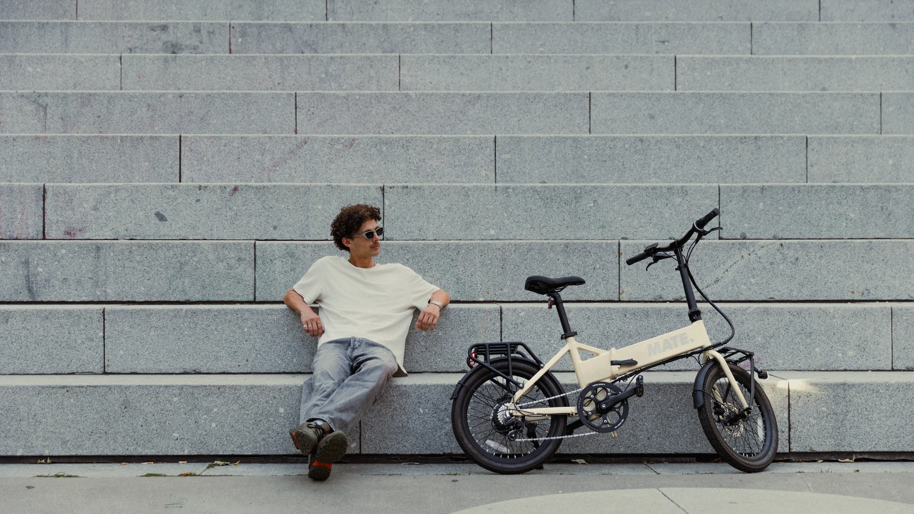 MATE GO kaufen: Beigefarbenes Faltrad E-Bike vor einer großen Steintreppe mit einem entspannt sitzenden jungen Mann mit Sonnenbrille.
