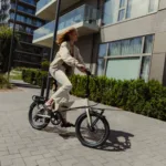 MATE GO kaufen: Eine junge Frau fährt auf einem beigefarbenen Faltrad E-Bike auf einem gepflasterten Weg vor modernen Wohngebäuden.
