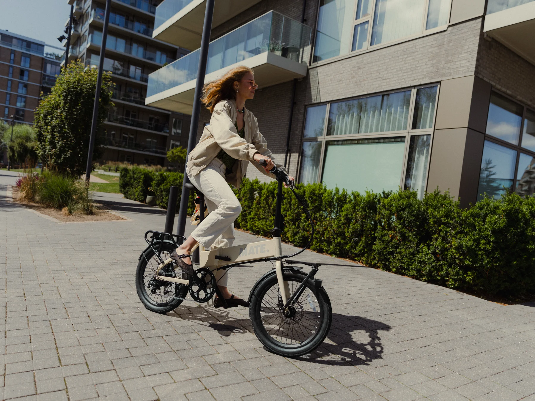 MATE GO kaufen: Eine junge Frau fährt auf einem beigefarbenen Faltrad E-Bike auf einem gepflasterten Weg vor modernen Wohngebäuden.