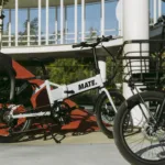 MATE GO+ kaufen: Weißes Faltrad E-Bike mit schwarzem Kindersitz auf dem Spielplatz für die Familie.