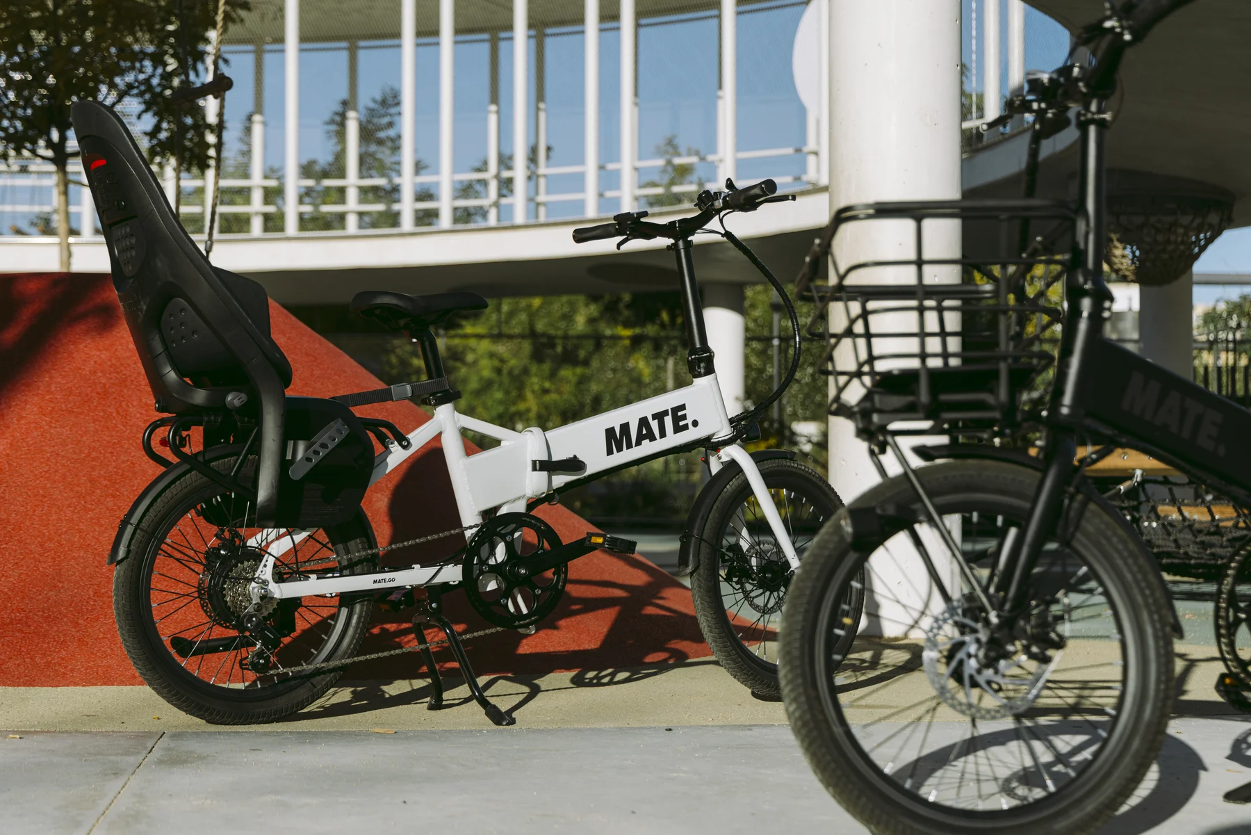MATE GO+ kaufen: Weißes Faltrad E-Bike mit schwarzem Kindersitz auf dem Spielplatz für die Familie.