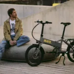 MATE GO+ kaufen: Graues Faltrad E-Bike in moderner Stadtumgebung mit Fahrer für urbane Pendler.