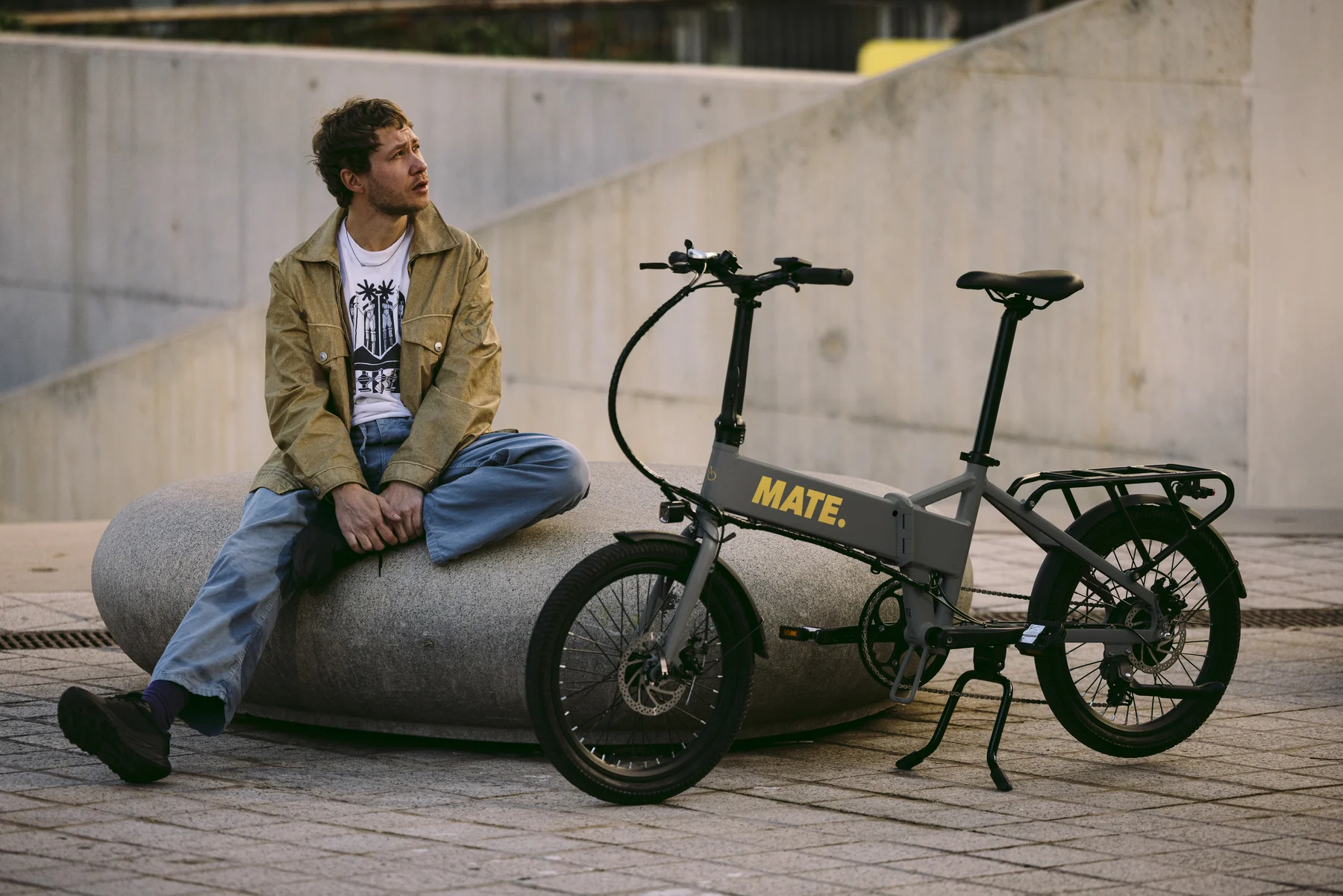 MATE GO+ kaufen: Graues Faltrad E-Bike in moderner Stadtumgebung mit Fahrer für urbane Pendler.