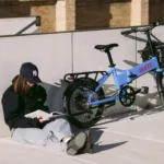 MATE GO+ kaufen: Hellblaues Faltrad E-Bike mit vorderem Gepäckkorb neben einer lesenden Frau an einer weißen Betonwand.