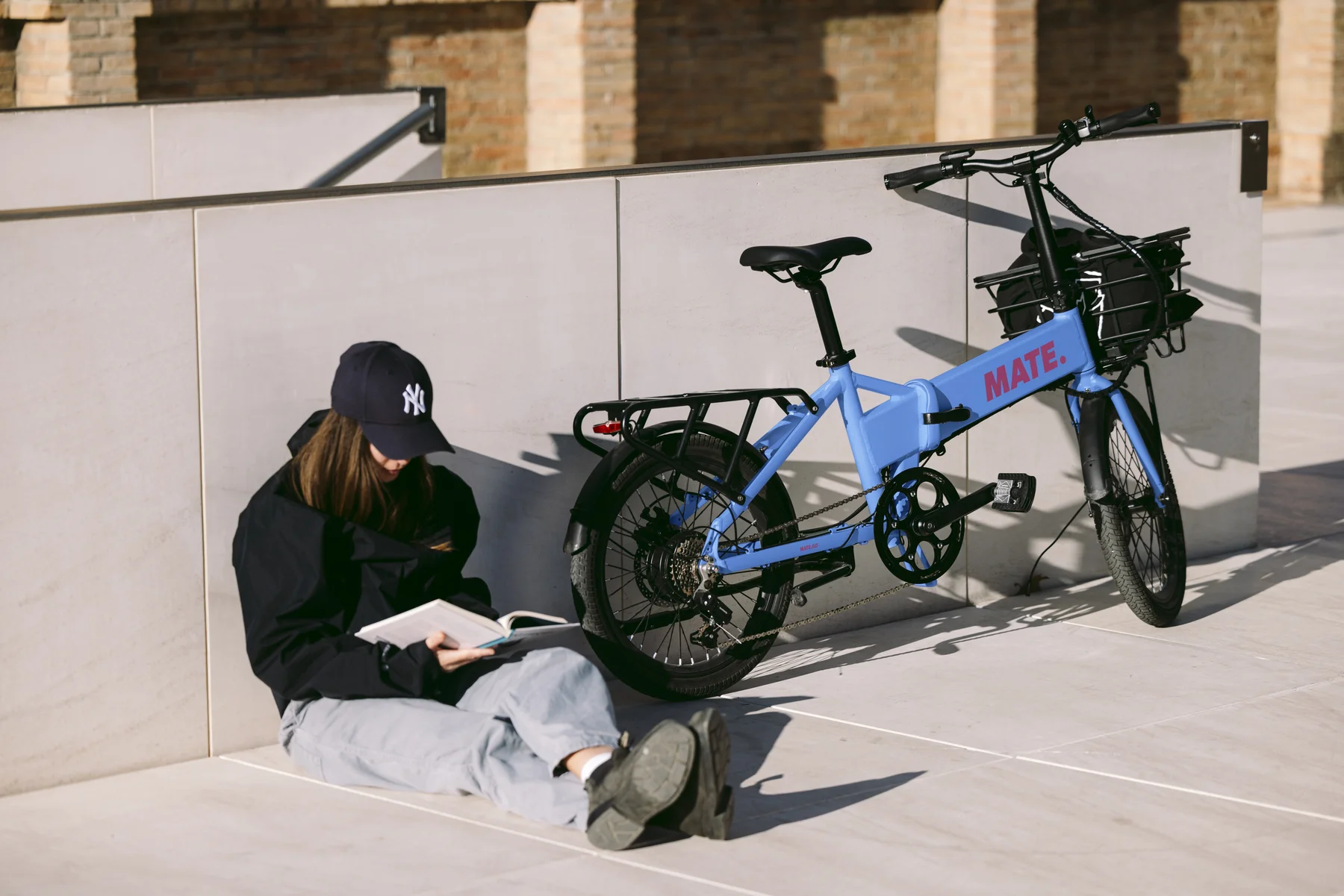 MATE GO+ kaufen: Hellblaues Faltrad E-Bike mit vorderem Gepäckkorb neben einer lesenden Frau an einer weißen Betonwand.