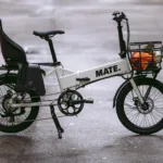 MATE GO+ kaufen: Weißes Faltrad E-Bike mit gefülltem Einkaufskorb vorne und schwarzem Kindersitz auf einer Asphaltstraße.