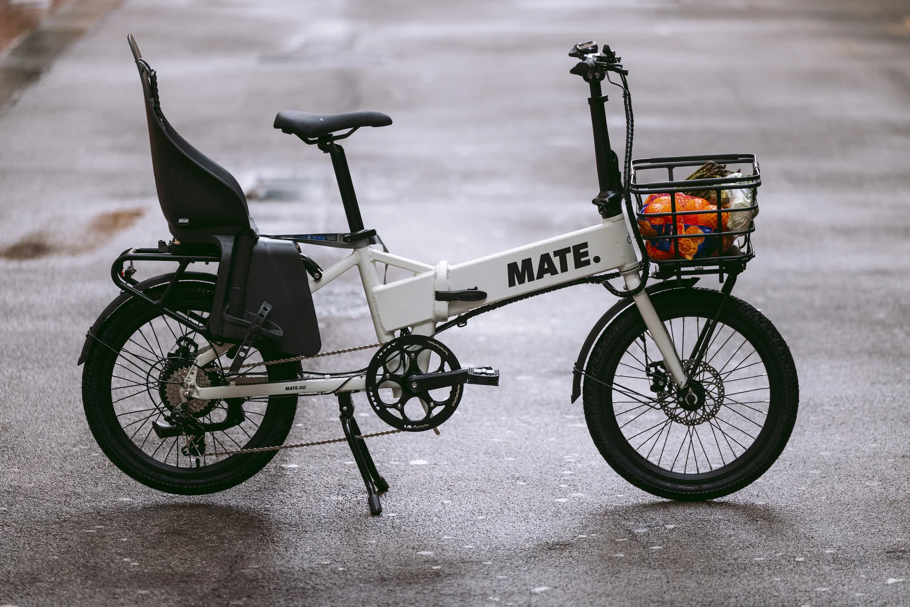 MATE GO+ kaufen: Weißes Faltrad E-Bike mit gefülltem Einkaufskorb vorne und schwarzem Kindersitz auf einer Asphaltstraße.