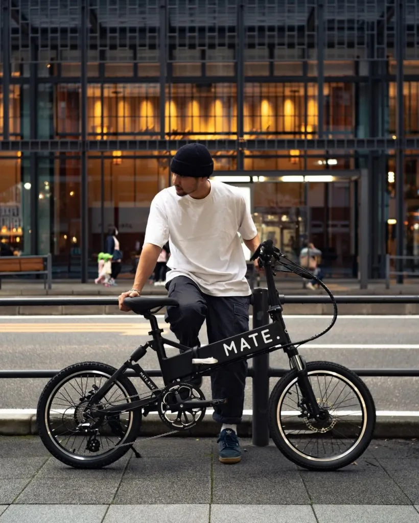 Urbanes MATE E-Klapprad in Schwarz mit einem Fahrradfahrer am Straßenrand vor einer gläsernen Fassade am Abend.