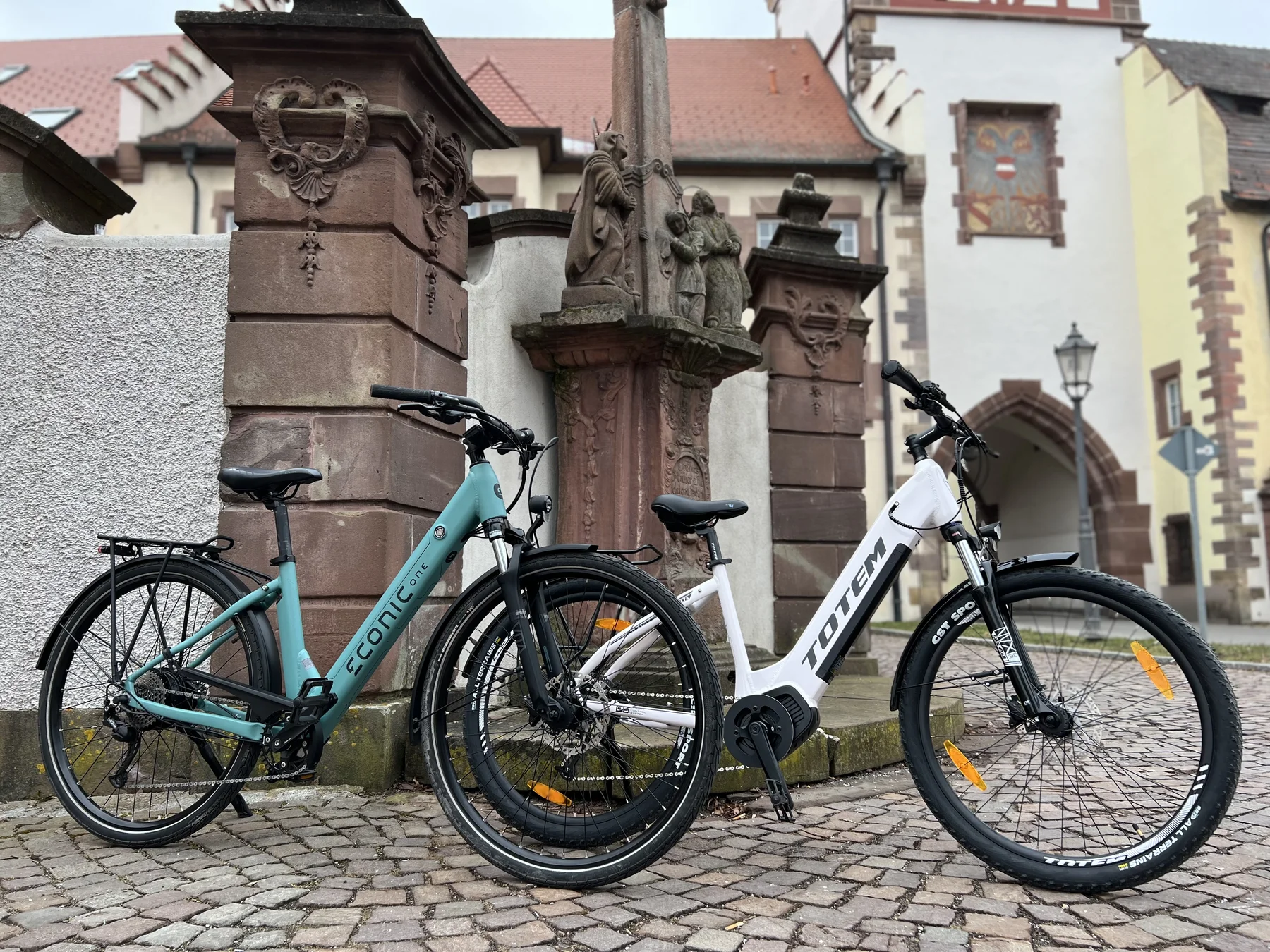 E-Bikes kaufen – Ein türkisfarbenes Econic One Tiefeinsteiger-E-Bike und ein weißes Totem Elektrofahrrad mit CST Sport Reifen geparkt auf Kopfsteinpflaster vor einer historischen Steinstatue und alten Gebäuden.
