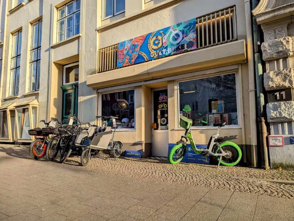 E-Bikes kaufen in Lübeck – cult.bike Lübeck GmbH: Die helle Ladenfront des Fahrradgeschäfts mit bunten Werbebannern und ausgestellten E-Bikes auf dem Gehweg davor.