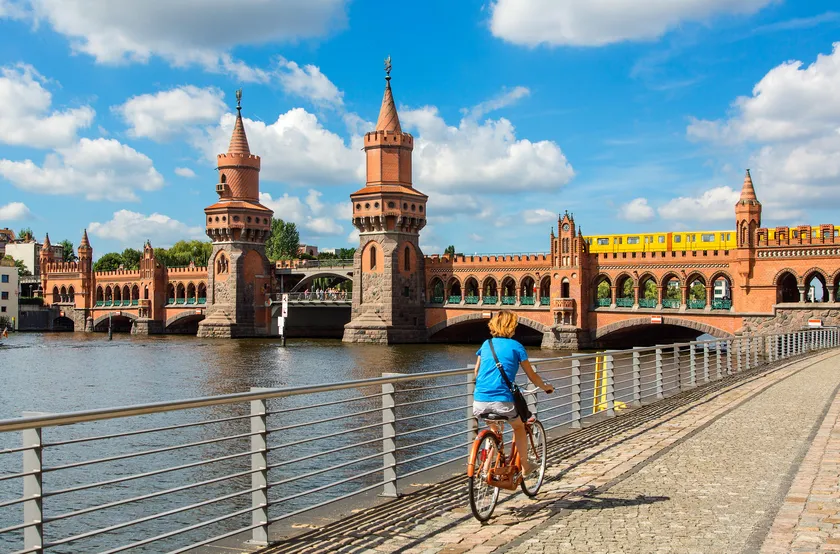 E-Bikes kaufen in Berlin-Pankow: Eine Fahrradfahrerin fährt auf der Uferpromenade mit Blick auf die historische Oberbaumbrücke und eine gelbe U-Bahn.