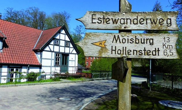 E-Bikes kaufen in Kakenstorf: Ein hölzerner Wegweiser am Estewanderweg mit Richtungsangaben nach Moisburg und Hollenstedt vor einem historischen Fachwerkhaus.