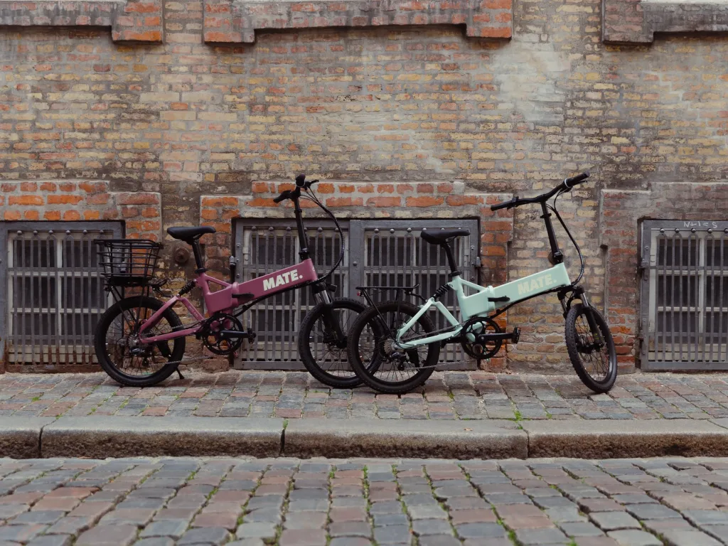 MATE City E-Bike kaufen bei E-Move24: Stilvolle MATE E-Klappräder in Soft Bloom und Marina Breeze abgestellt an einer gepflasterten Straße vor einer alten Ziegelmauer.