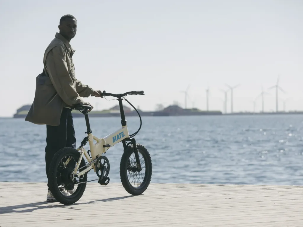 MATE FUSION E-Bike kaufen bei E-Move24: Elegantes MATE E-Klapprad in Beige am Wasser mit einem Mann auf einer Holzpromenade und Windrädern im Hintergrund.