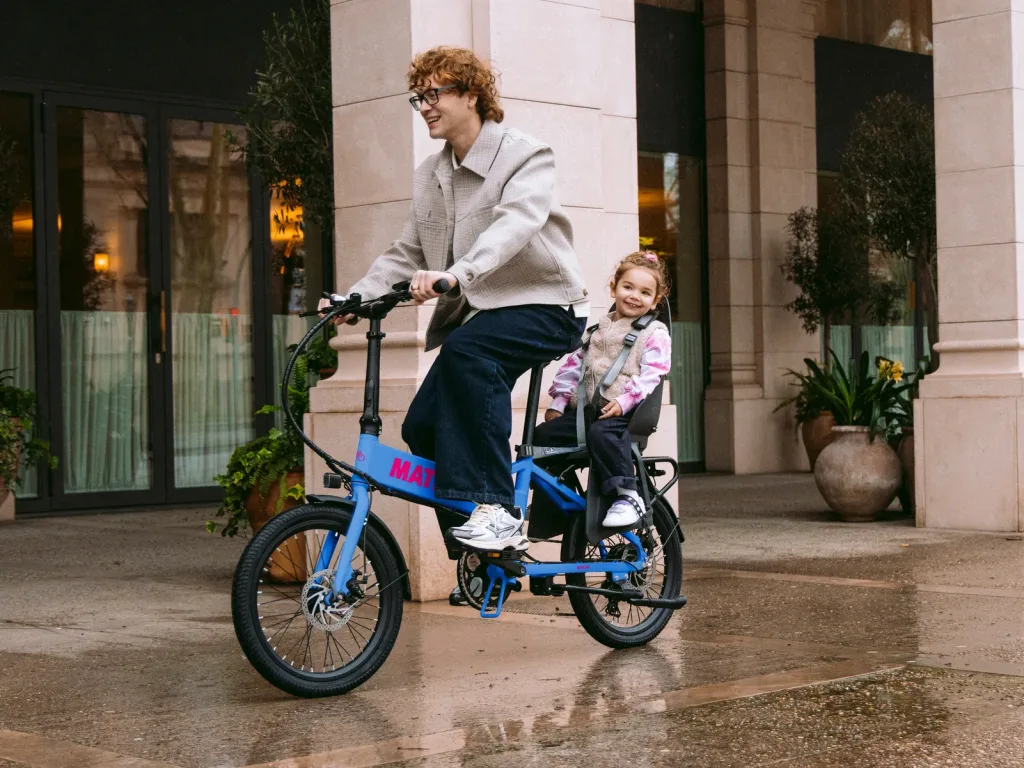 Blaues MATE GO Cargo-E-Bike mit montiertem Kindersitz auf dem Gepäckträger bei einer Fahrt mit Vater und Tochter in der Stadt.