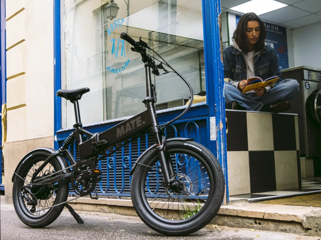 MATE X EVO E-Bike kaufen bei E-Move24: Urbanes MATE E-Klapprad in Schwarz abgestellt vor einer Wäscherei mit einer lesenden Person im Hintergrund.