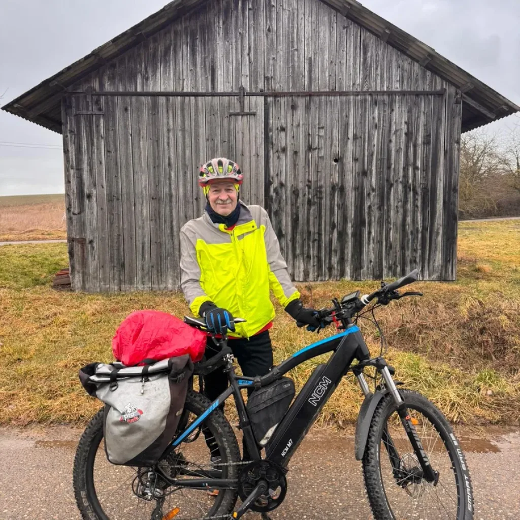 Echte Menschen. Echte Erfahrungen – Ein Mann steht vor einem E-Bike Lagerhäuschen mit seinem modernen E-Bike